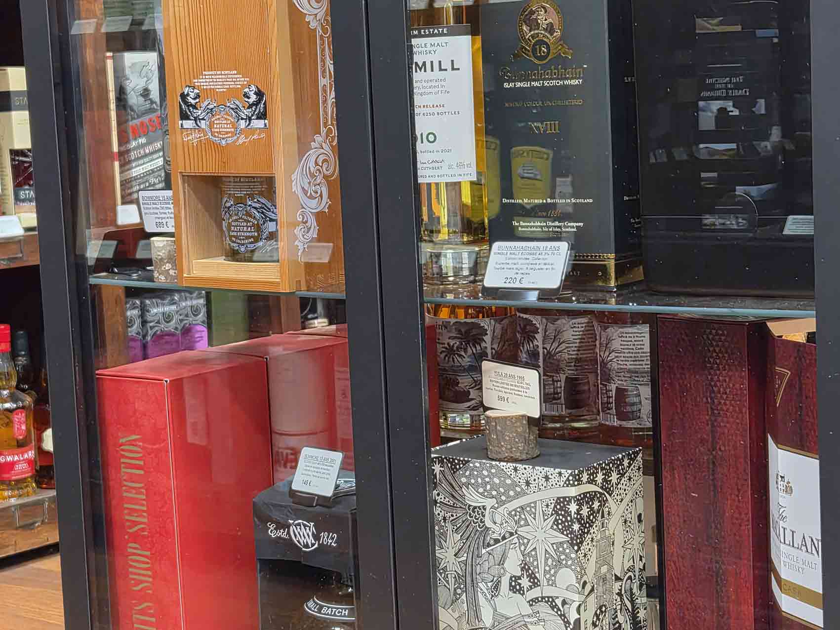 Whiskies rares : pourquoi certaines bouteilles deviennent-elles si recherchées ? 3 Vitrine en verre exposant des bouteilles de whisky rares et chères. On y voit un Bunnahabhain 18 ans, un Yula 20 ans, un coffret Bowmore, un The Macallan rare collection 2021 release et un Daftmill.