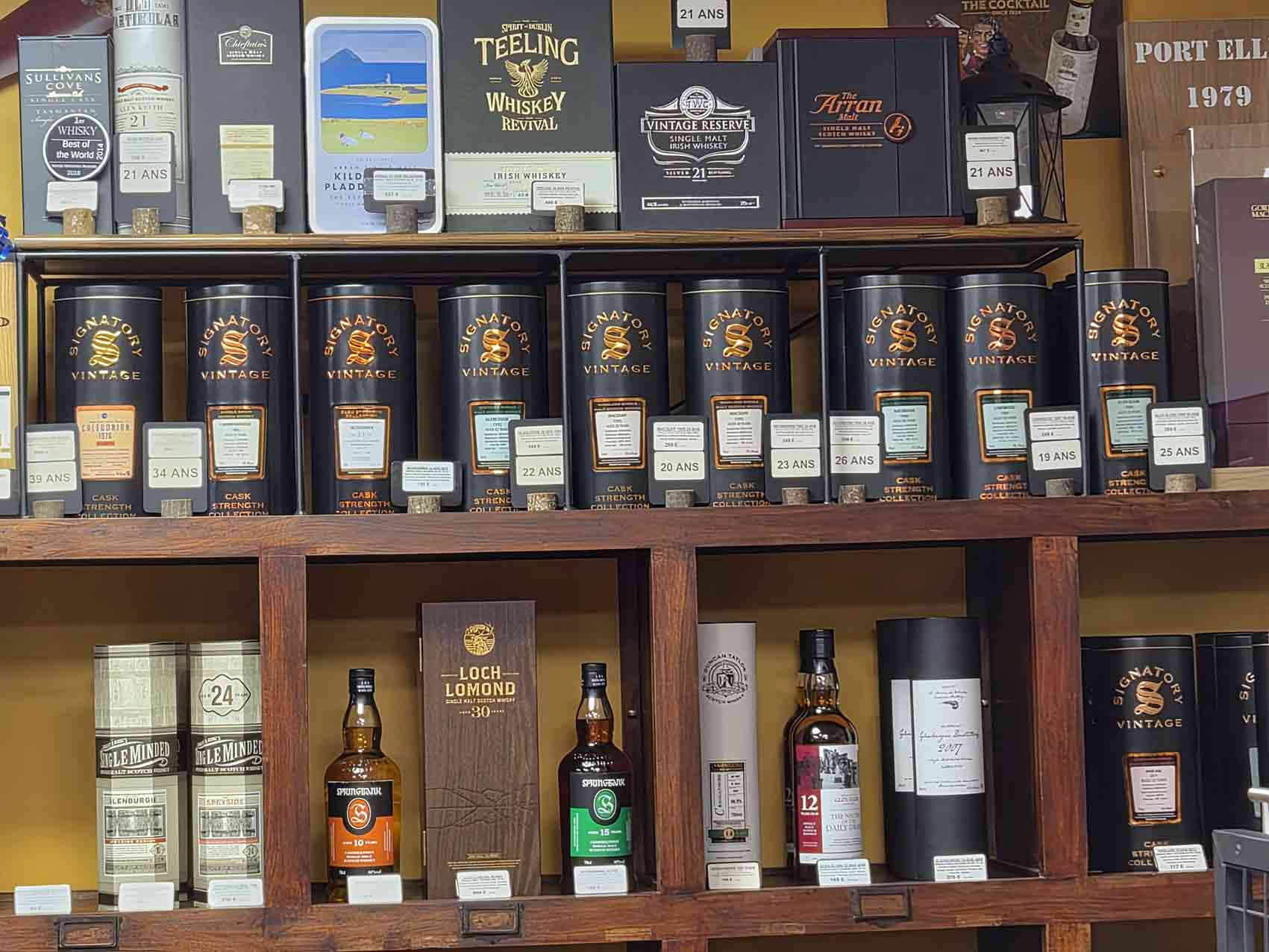 Whiskies rares : pourquoi certaines bouteilles deviennent-elles si recherchées ? 2 Étagères en bois garnies de flacons et coffrets de whiskies d'exception, incluant des embouteillages indépendants Signatory Vintage, Sullivans Cove, Teeling Revival, Loch Lomond 30 ans et Springbank.